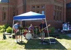 Photo of three men playing musical instruments under a blue canopy in front of a brick building